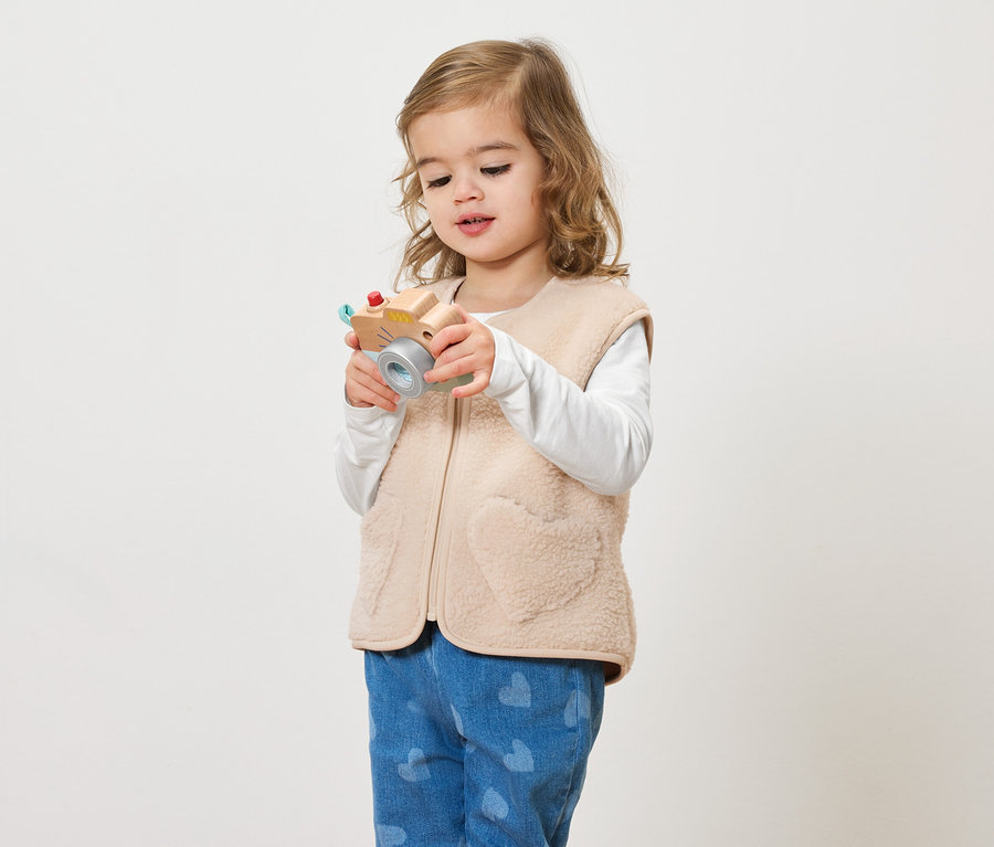 Ein kleines Mädchen mit lockigem Haar hält eine Spielzeugkamera vor einem hellen Hintergrund.