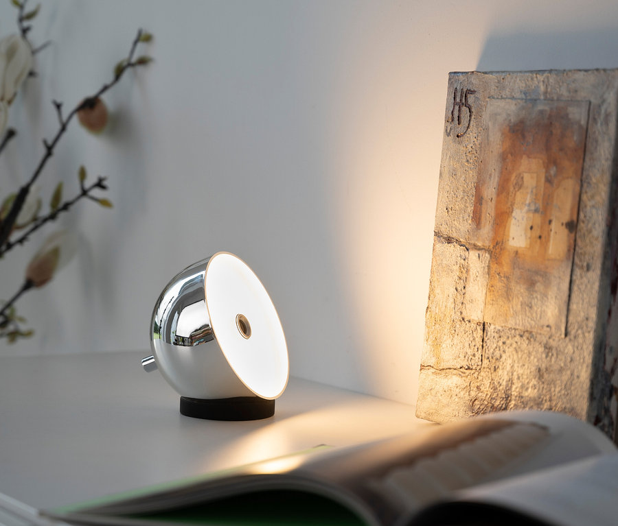 Silberfarbene SCHÖNER WOHNEN Akku-Tischleuchte »Bellis« leuchtet auf einem Tisch neben einem Buch und einem Dekostück.