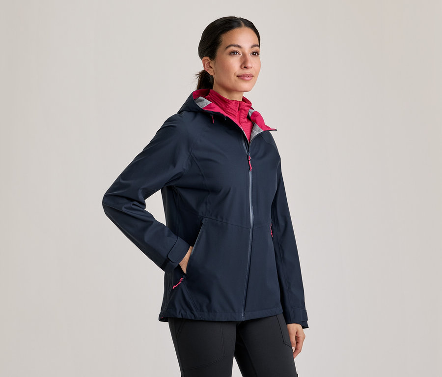 Frau trägt eine dunkelblaue Jacke mit pinkem Innenfutter und schwarzen Hosen, die Hand in der Tasche.