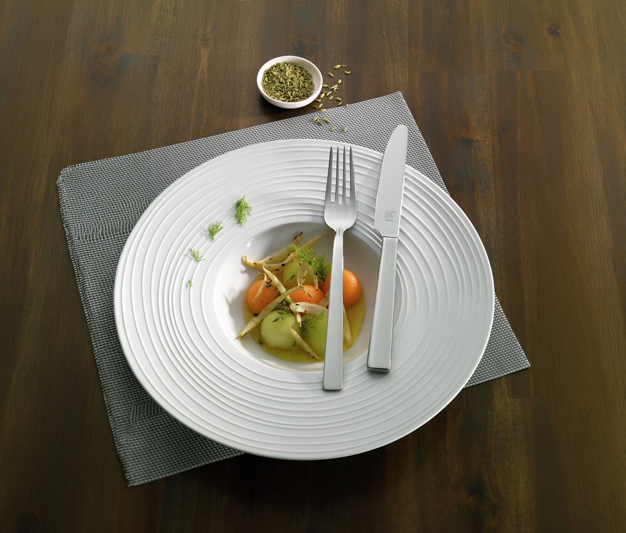 Weißer Teller mit Melonen-Salat, Gabel und Messer des ZWILLING »King« Besteck-Sets auf grauem Platzset. Daneben Schale mit Gewürzen auf Holztisch.