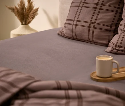 Gemütliches Bett mit grauem Spannbettlaken, zwei karierten Kissen und passender Bettdecke. Eine weiße Tasse mit Getränk steht auf einem Holztablett auf dem Bett. Im Hintergrund eine Vase mit getrocknetem Pampasgras.