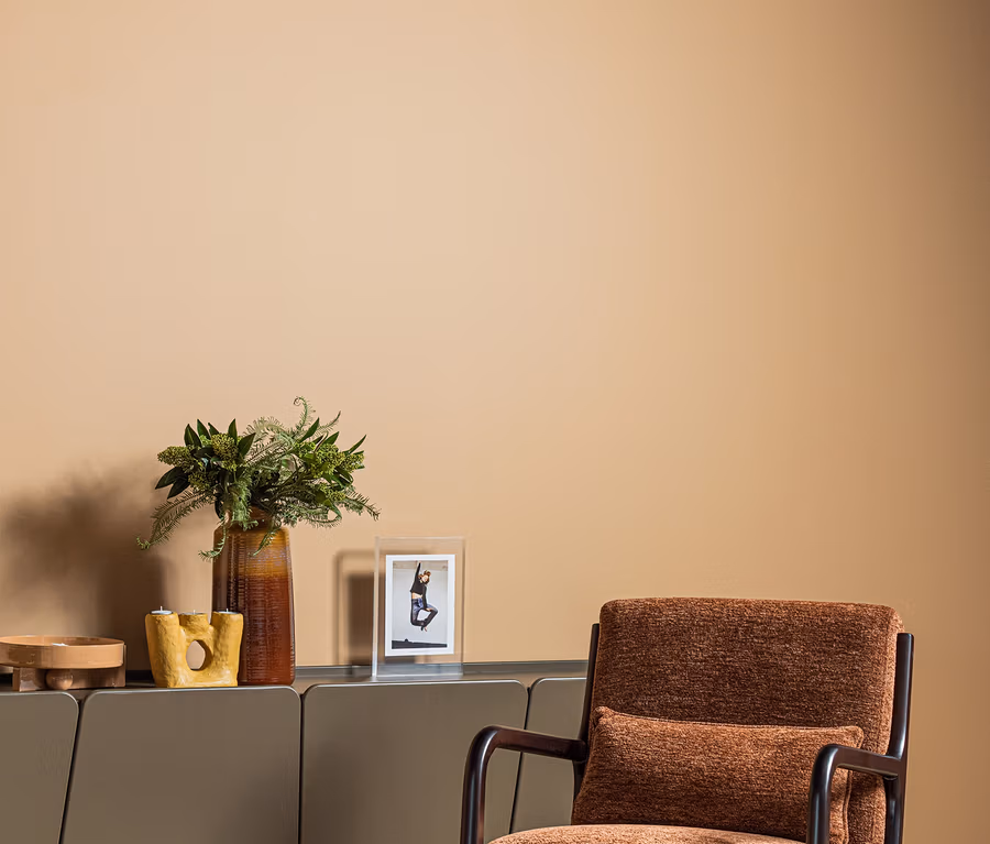 Ein brauner Polstersessel mit dunklen Holzarmlehnen und -beinen steht auf einem hellen Holzboden. Dahinter steht ein graues Sideboard, auf dem eine hohe braune Vase mit grünem Blattwerk, ein gelber abstrakter Kerzenhalter, eine kleine Holzschale und ein gerahmtes Bild einer Person stehen. Die Wand dahinter ist einfarbig beige.