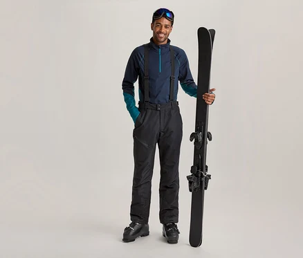Lächelnder Mann in dunkelblauer Skibekleidung mit Hosenträgern und Skibrille auf der Stirn, der schwarze Skier aufrecht hält.