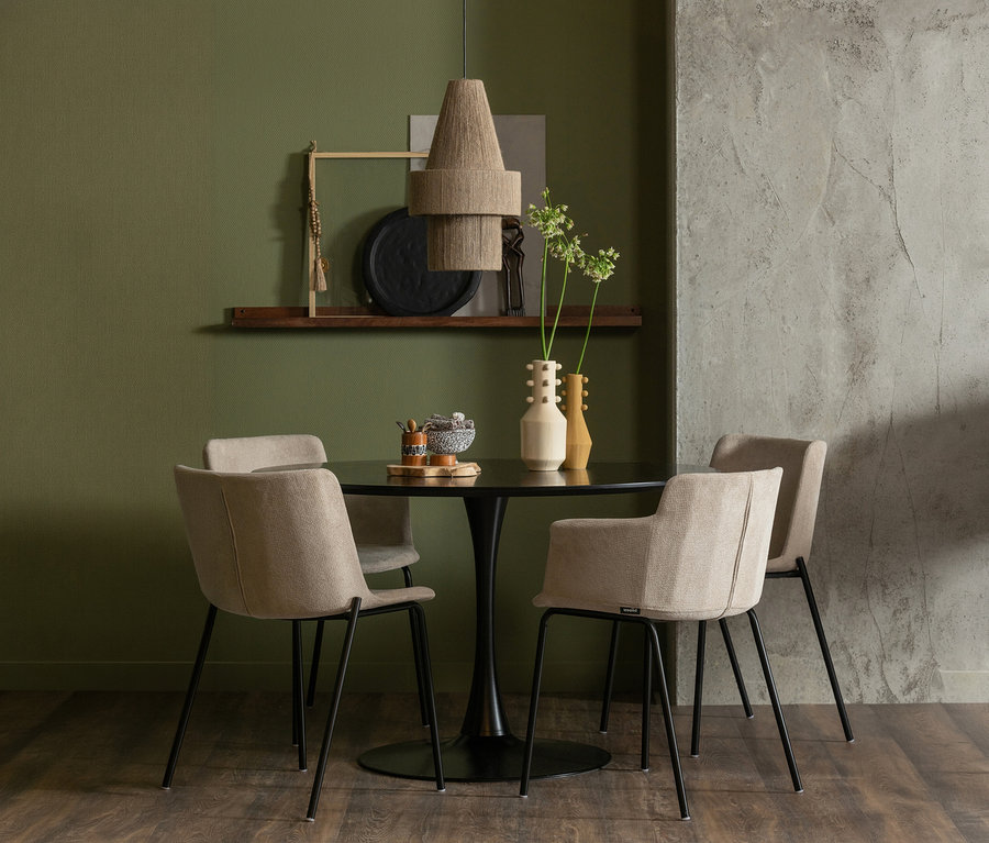 Ein Essbereich mit einem runden schwarzen Tisch und vier beigefarbenen Polsterstühlen auf dunklem Holzboden. Eine gestufte, gewebte Pendelleuchte hängt über dem Tisch, der mit zwei Vasen und einem kleinen Tablett dekoriert ist. Der Raum hat eine olivgrüne Wand mit einem Regal und eine strukturierte graue Betonwand.