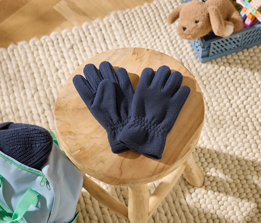 Dunkelblaue Fleecehandschuhe liegen auf einem Holzhocker. Daneben eine hellblaue Tasche mit einer dunkelblauen Mütze. Im Hintergrund ein Teddybär in einer blauen Kiste auf einem Teppich.
