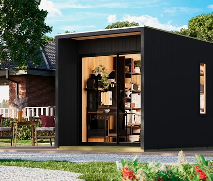 Ein modernes, schwarzes Gartenbüro mit einer großen Glasschiebetür, die einen Innenraum mit Regalen und einem Schreibtisch zeigt. Links daneben befindet sich eine Terrasse mit zwei Korbsesseln mit roten Polstern und einem kleinen Tisch. Im Hintergrund ist ein Backsteinhaus zu sehen, im Vordergrund ein Kiesweg und Blumen.