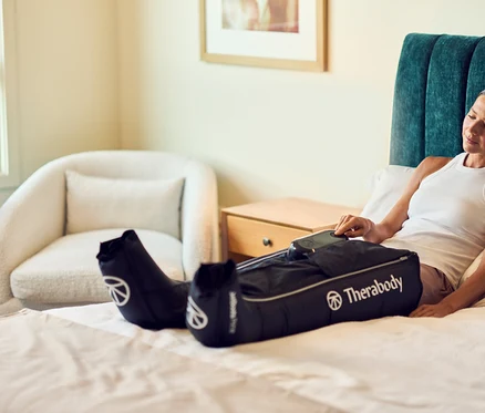 Eine Frau entspannt sich auf einem Bett mit geschlossenen Augen, trägt schwarze Therabody Kompressionsstiefel an den Beinen und bedient ein Steuergerät.