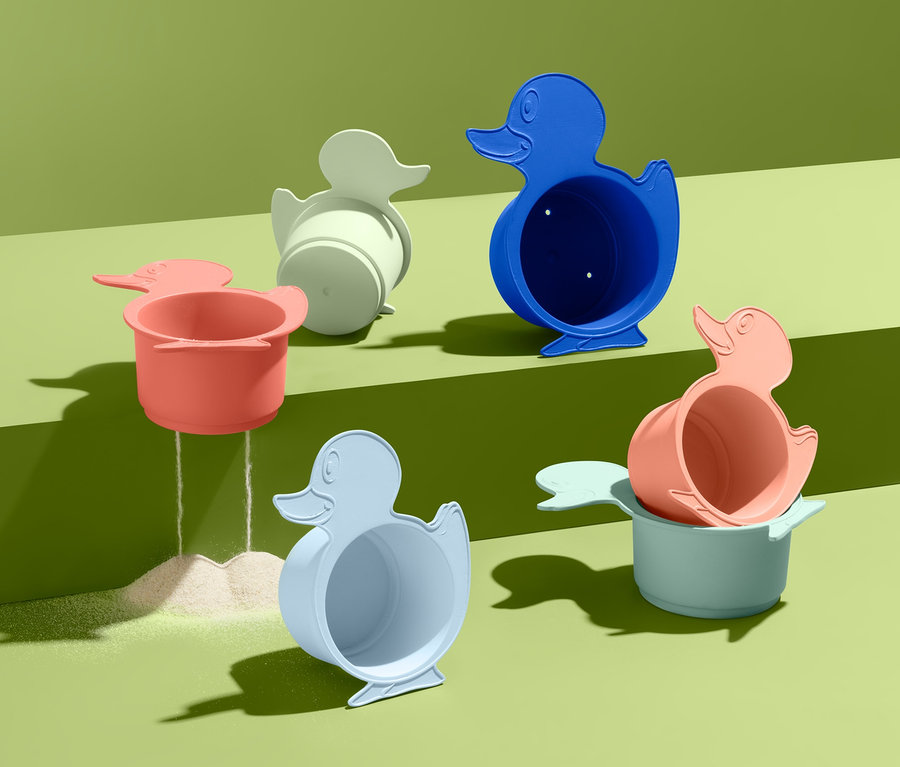 Sechs bunte Bade- und Sandspielbecher in Entenform stehen auf grünem Grund, aus einem rieselt Sand.