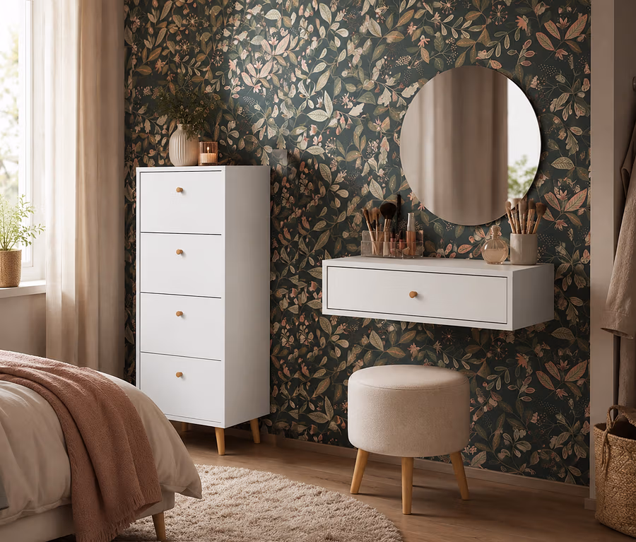 Ein Schlafzimmer oder Ankleidebereich mit dunkler Blumentapete. Links steht eine matte weiße Kommode »Bjarne« mit 4 Schubladen neben einem Bett. Rechts hängt ein runder Spiegel über einem schwebenden weißen Waschtisch mit Make-up, darunter ein beiger Hocker.