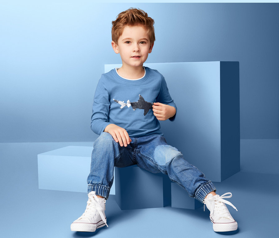 Junge sitzt auf einem blauen Würfel und trägt ein blaues Langarmshirt mit Hai-Motiv, Jeans und weiße Turnschuhe.
