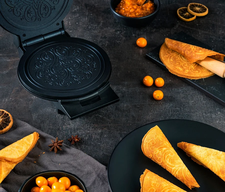 Schwarzes WILFA Norwegisches Krumkakeeisen ØSTERDAL Waffeleisen mit gebackenen Krumkaken, einer Schale Obst und Marmelade auf dunklem Hintergrund.