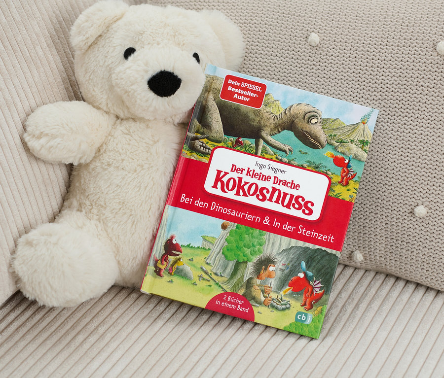 Ein Teddybär liegt neben dem Buch "Der kleine Drache Kokosnuss ‒ bei den Dinosauriern & In der Steinzeit".