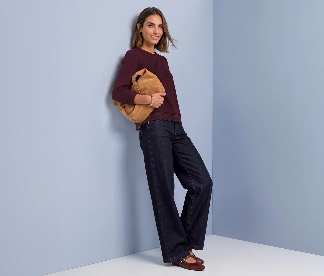 Lächelnde Frau in rotem Street One 3/4-Jerseyshirt mit Wellenkante, weiten Jeans, hält braune Tasche, an blauer Wand lehnend.