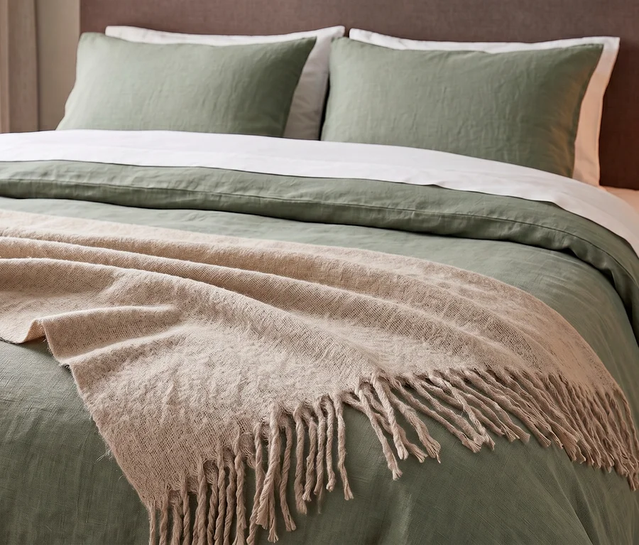 Ein Bett mit salbeigrüner Bettwäsche und zwei Kissen. Eine beige SCHÖNER WOHNEN-Kollektion Wohndecke »PLENTY« mit Fransen liegt über dem Fußende des Bettes.