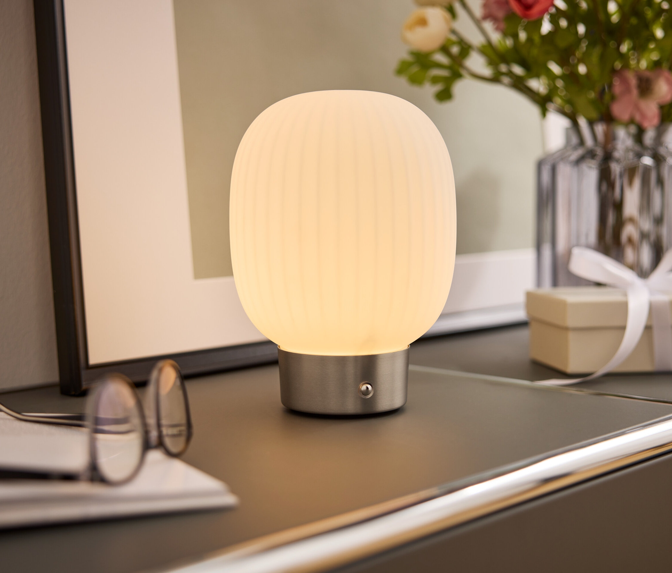 Eine Nachttischlampe mit weißem Schirm steht auf einem Schrank neben einer Brille, einem Buch, einer Blumenvase und einer Geschenkbox.