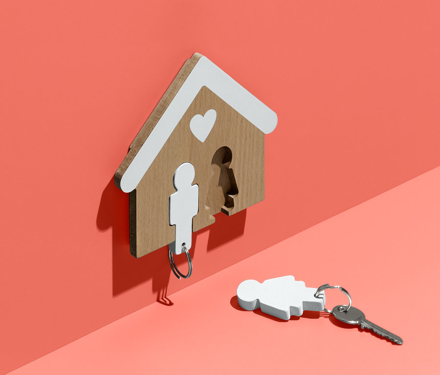 An einer korallenroten Wand hängt ein hölzernes Schlüsselbrett in Form eines Hauses mit weißem Dach und Herz. Ein Schlüssel mit einem Figurenanhänger hängt am Brett, ein weiterer liegt am Boden.
