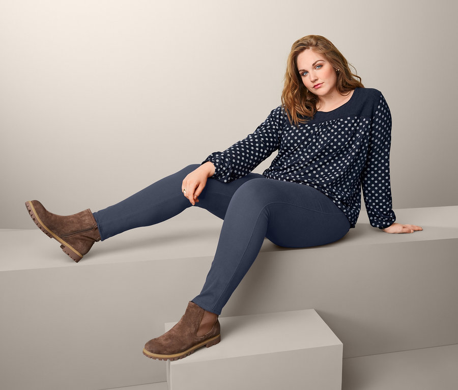 Frau sitzt auf grauer Plattform und trägt Veloursleder-Boots, Blusenshirt mit Stickerei und Jeggings.