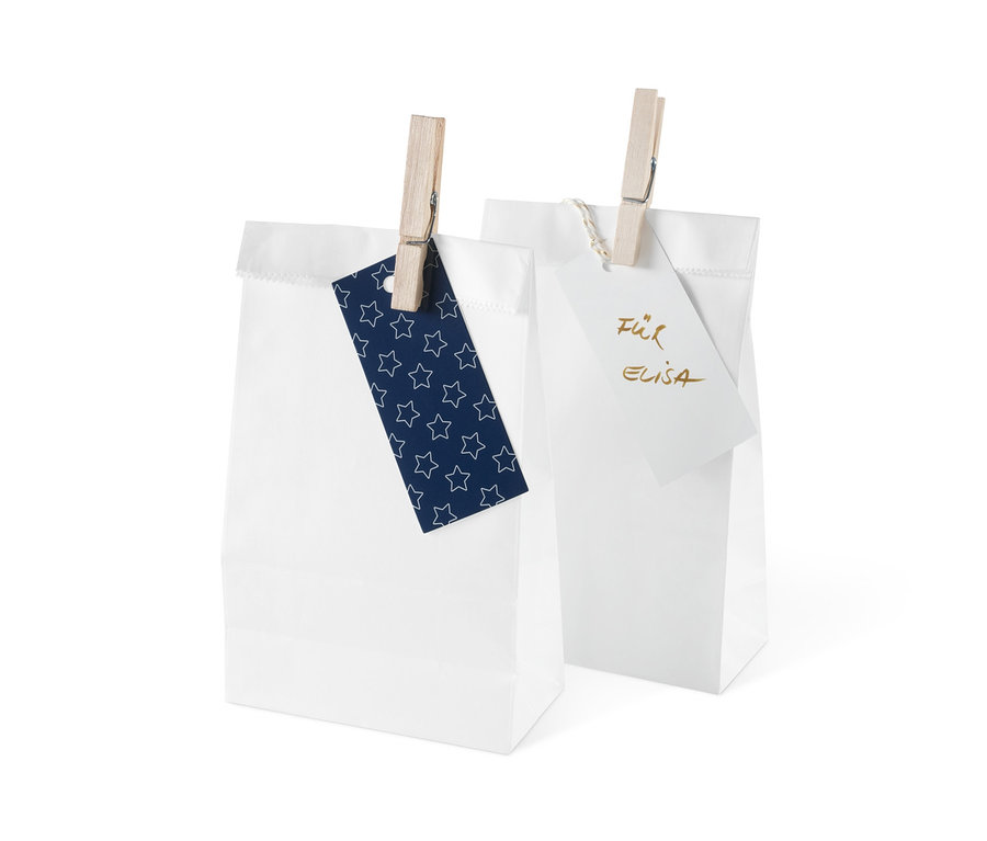 Zwei weiße Geschenktüten aus dem Adventskalender-und-Geschenktüten-Set sind mit einer Wäscheklammer verschlossen. An einem hängt ein blaues Schild mit Sternen, am anderen ein weißes Schild mit Schrift.
