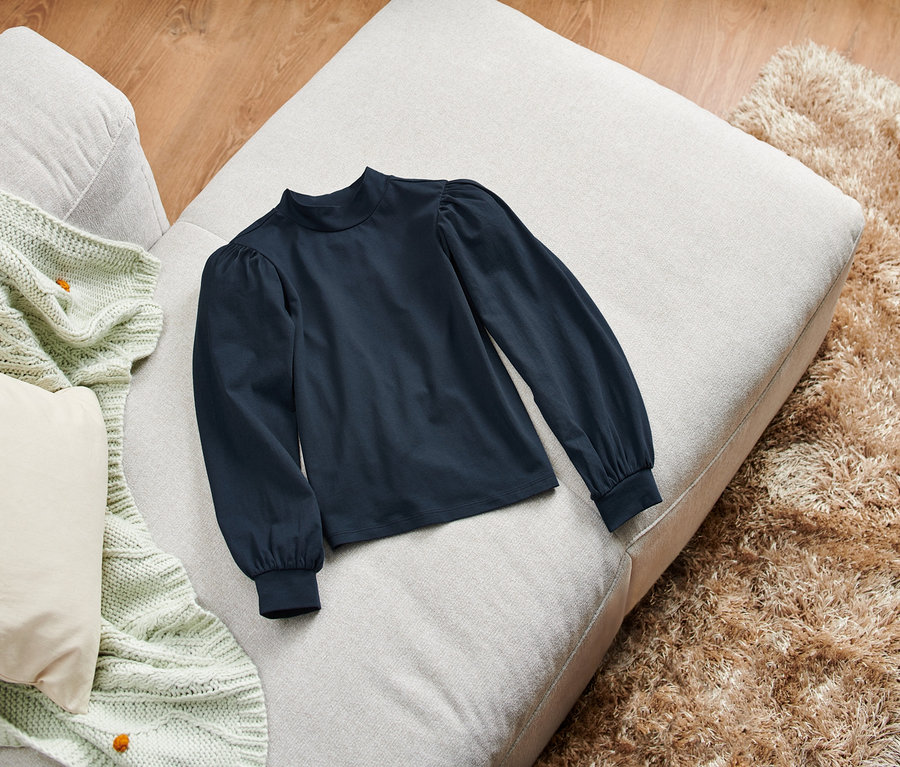 Ein dunkelblaues Langarmshirt liegt auf einem beigen Pouf neben einer hellgrünen Decke und einem zotteligen Teppich.