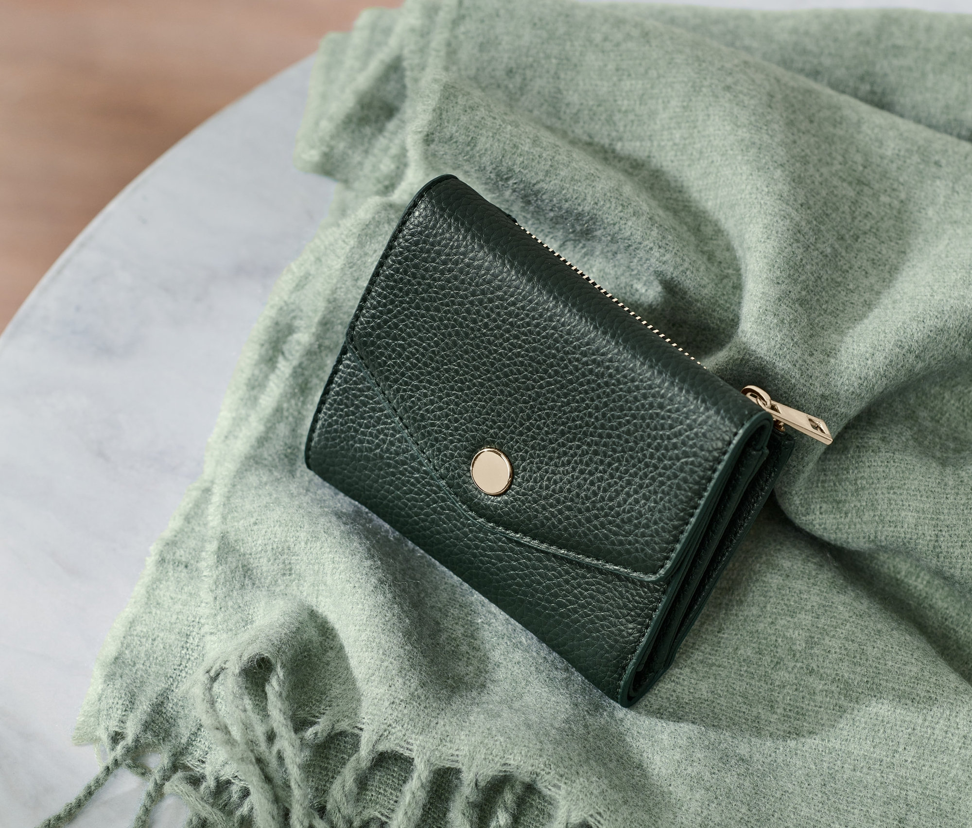 Eine kleine Geldbörse liegt auf einem grünen Schal mit Fransen auf einem Marmortisch.