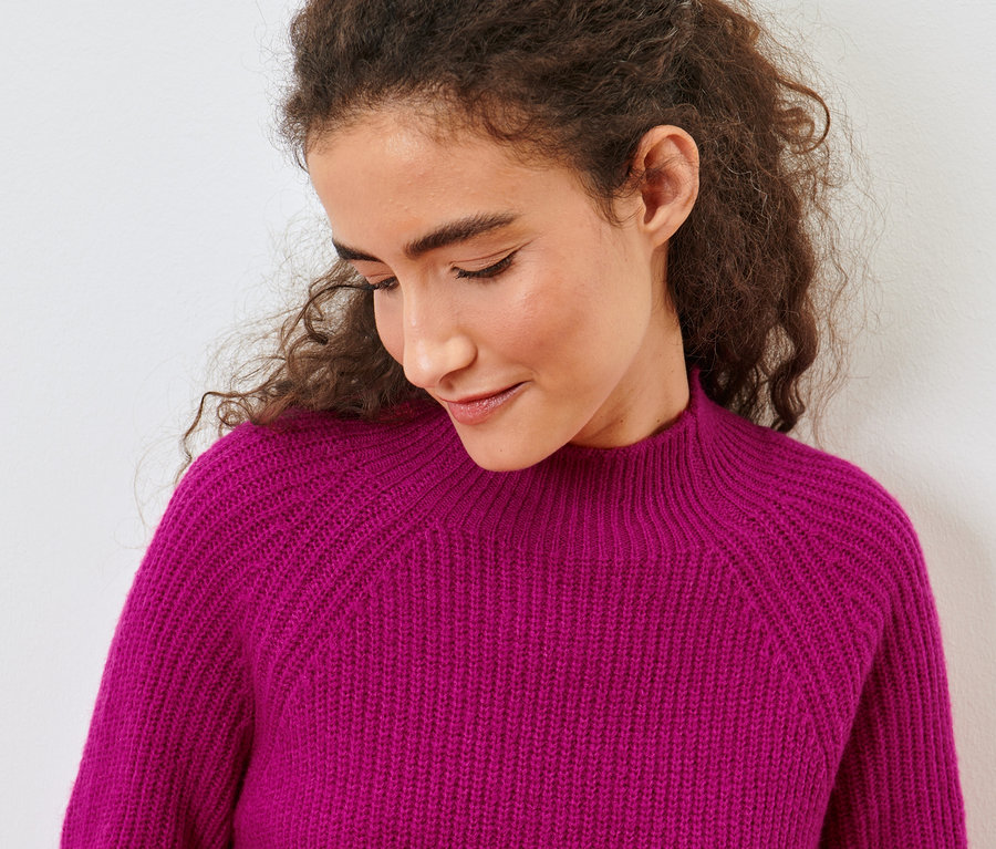Porträt einer Frau mit braunen lockigen Haaren, die einen pinkfarbenen Strickpullover mit Stehkragen trägt.