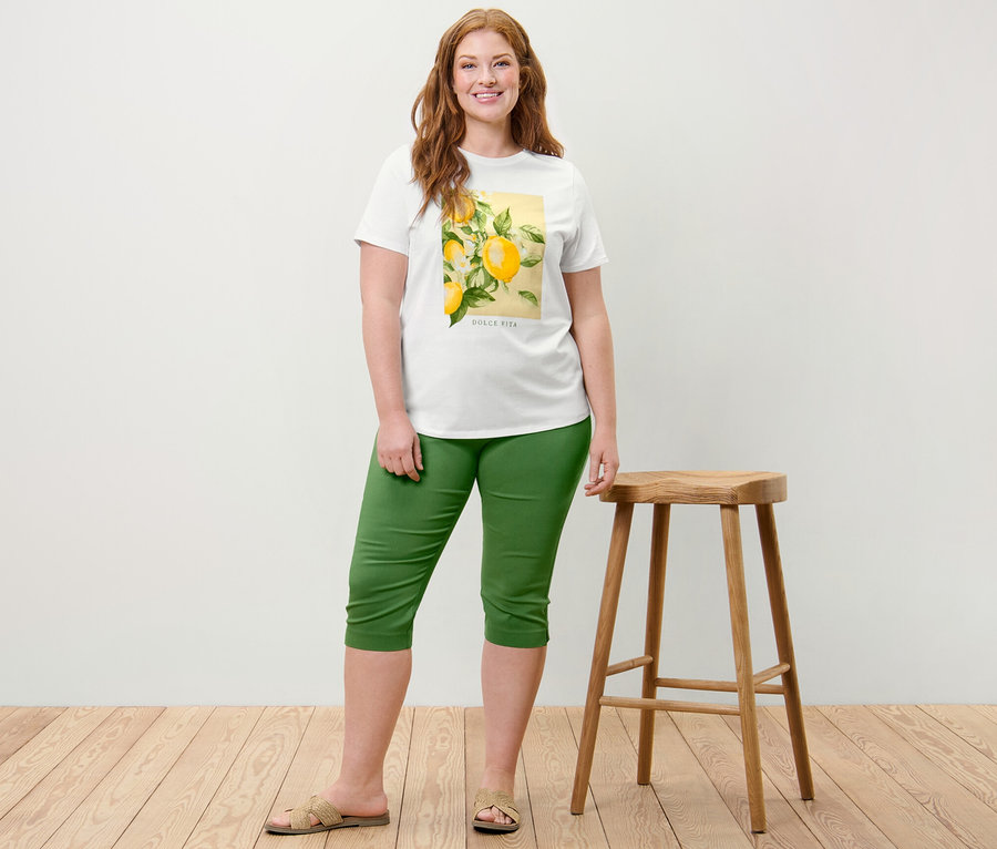 Frau posiert neben einem Holzhocker und trägt ein T-Shirt mit Print und eine grüne 3/4-Stretchhose.
