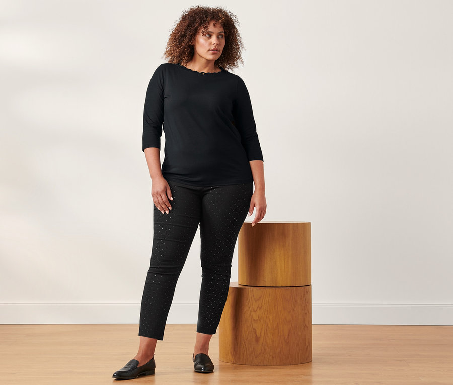 Eine Frau steht und trägt ein schwarzes Shirt mit 3/4-Arm und eine gepunktete Stretchhose.