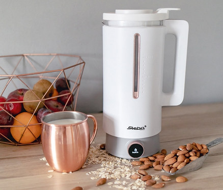 Auf einer Holztheke stehen ein weißer Vegan-Drink-Maker, ein Kupferbecher, ein Löffel Mandeln, Haferflocken und ein Korb mit Früchten. Zu sehen ist der Steba Vegan-Drink-Maker »VDM 2 HOT & COLD«.