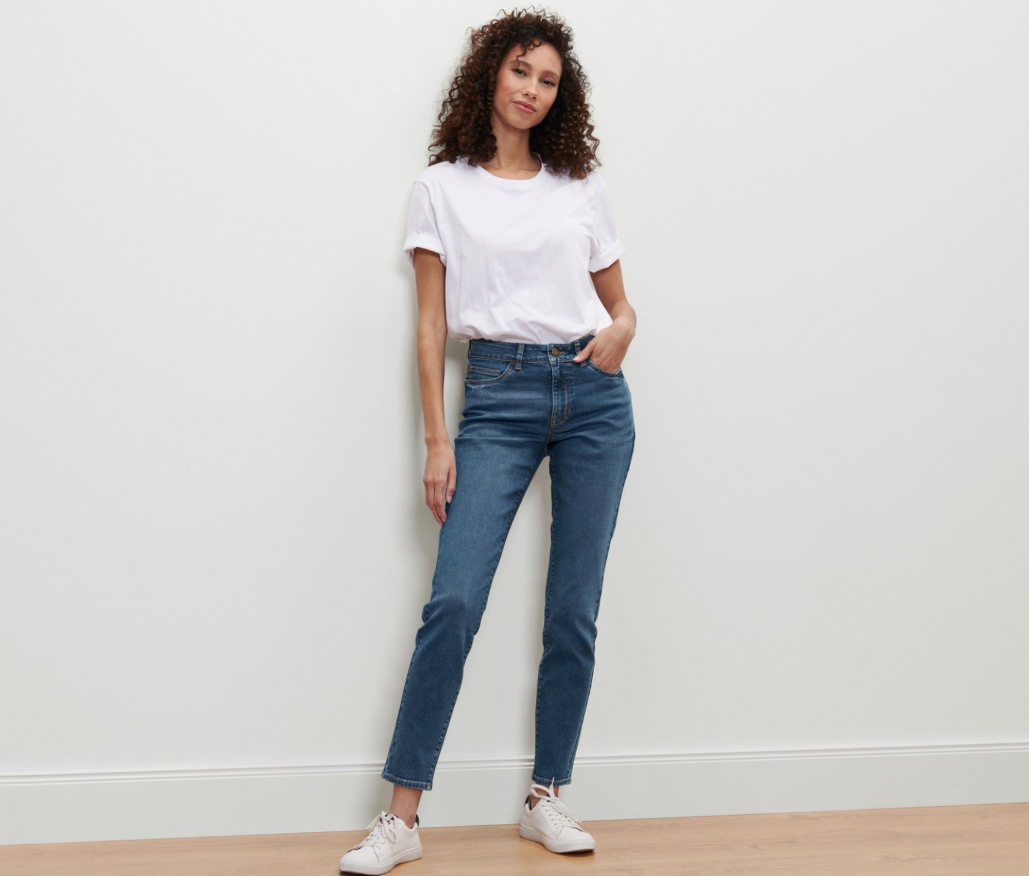 Frau lehnt an Wand und trägt Slim-Denim »Fit Emma«, midblue Denim, weißes T-Shirt und Turnschuhe.