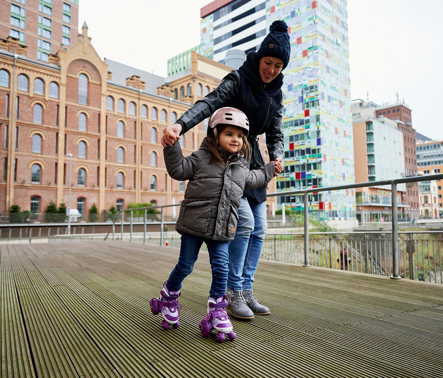 Eine Mutter hält die Hände ihrer Tochter, die lila HUDORA Rollschuhe "My First Quad" trägt.