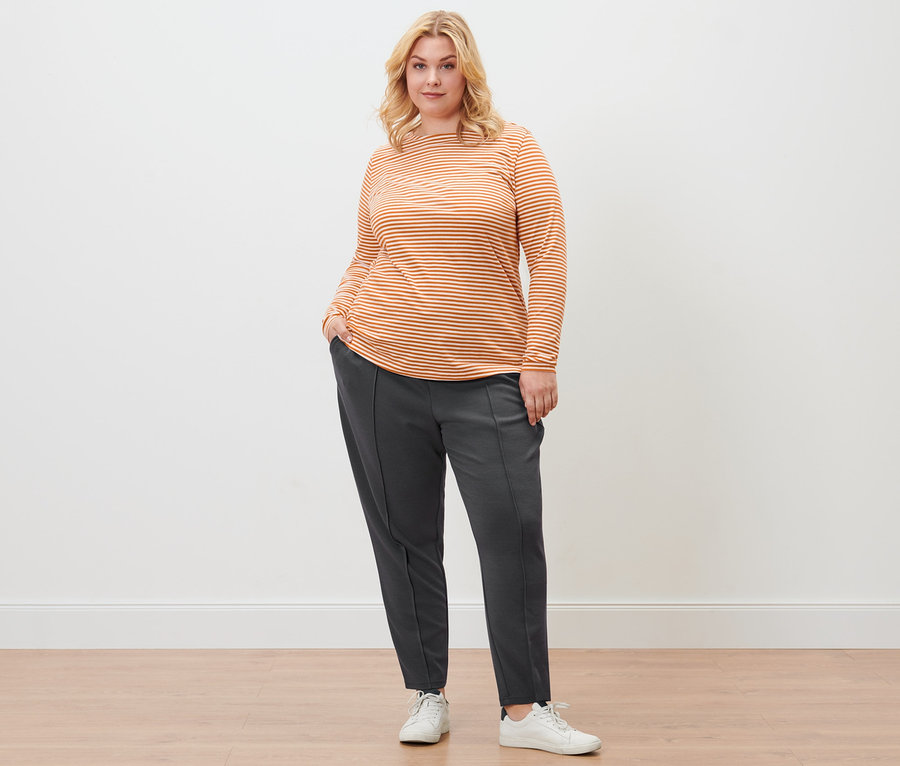 Eine Frau posiert in einem orangefarbenen, gestreiften Langarmshirt, einer Joggpants mit Biese und Ledersneakern.