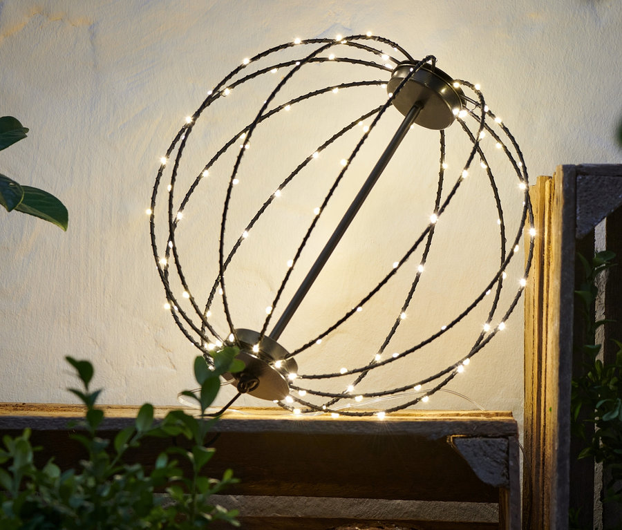 Eine LED-Kugel aus Metall steht auf einer Holzkiste neben Pflanzen.