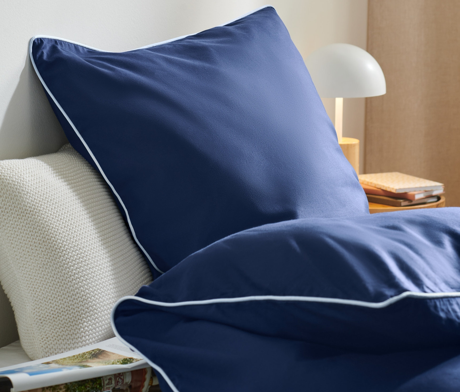 Detailansicht von marineblauer Baumwoll-Bettwäsche mit TENCEL™ Lyocell und weißer Paspel auf einem Bett.
