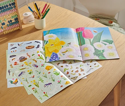 Auf einem Tisch liegen ein geöffnetes Buch "Hurra! Endlich Frühling", ein Abakus, ein Becher mit Buntstiften, ein Locher und Stickerbögen.