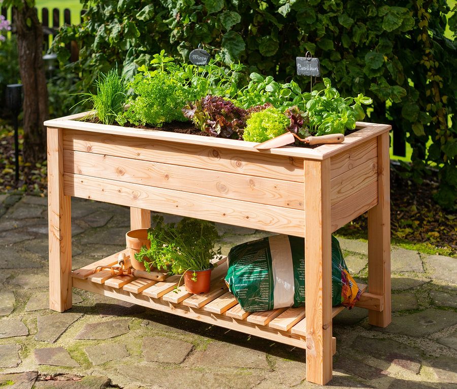 Ein hölzernes Stelzenhochbeet mit Pflanzen und Gartengeräten steht im Freien.