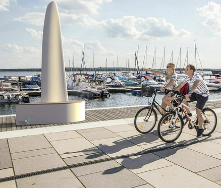 Zwei Personen fahren mit Fahrrädern auf einer Promenade am Wasser, im Hintergrund Boote und eine weiße Wings for Living Bank »Sydney«.