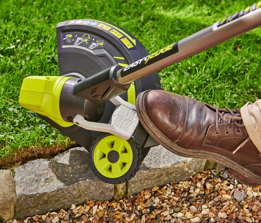 Detailaufnahme eines RYOBI 18 V One+ Akku-Rasentrimmers mit Radführung und einem Fuß in einem braunen Schuh, der ihn bedient.