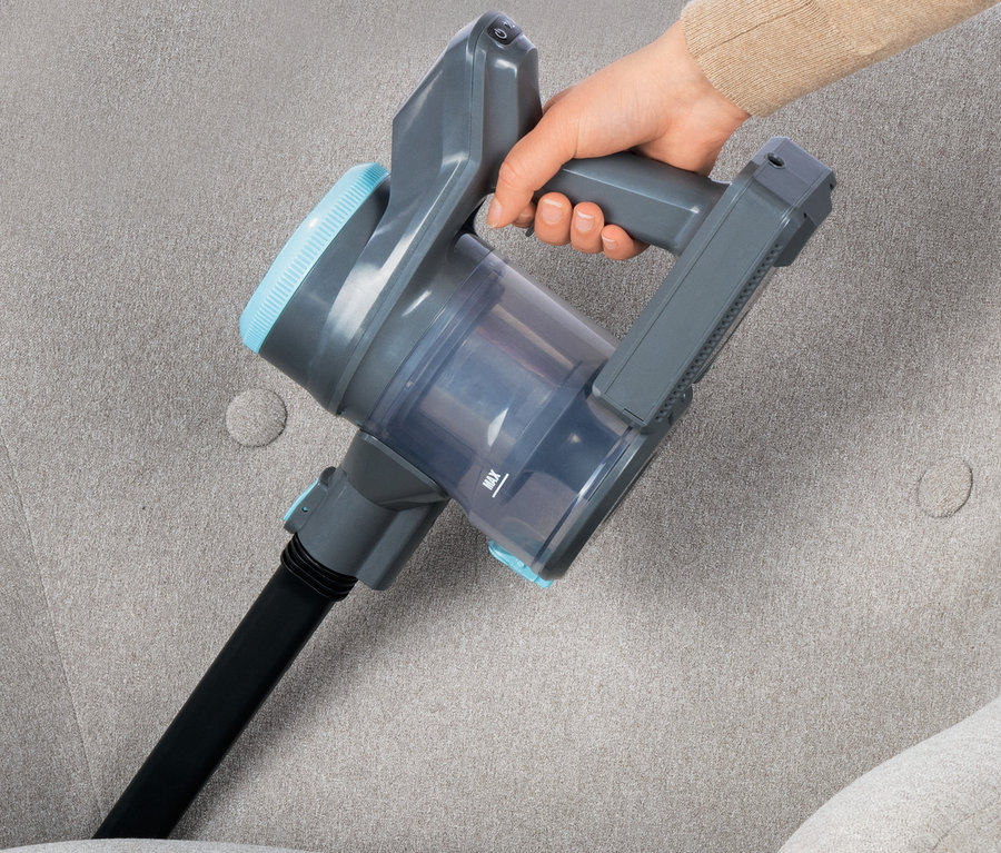 Eine Hand hält einen Akku-Hand- und Bodenstaubsauger auf einem grauen Sofa.