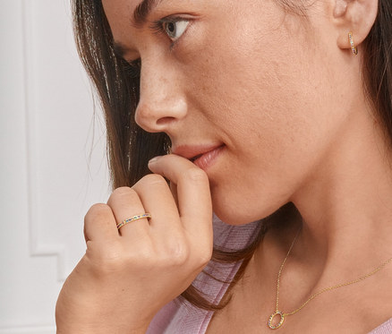 Profil einer brünetten Frau, die Gold-Creolen mit farbigen Zirkonia, eine Gold-Kette und einen Gold-Ring mit farbigen Zirkonia trägt.