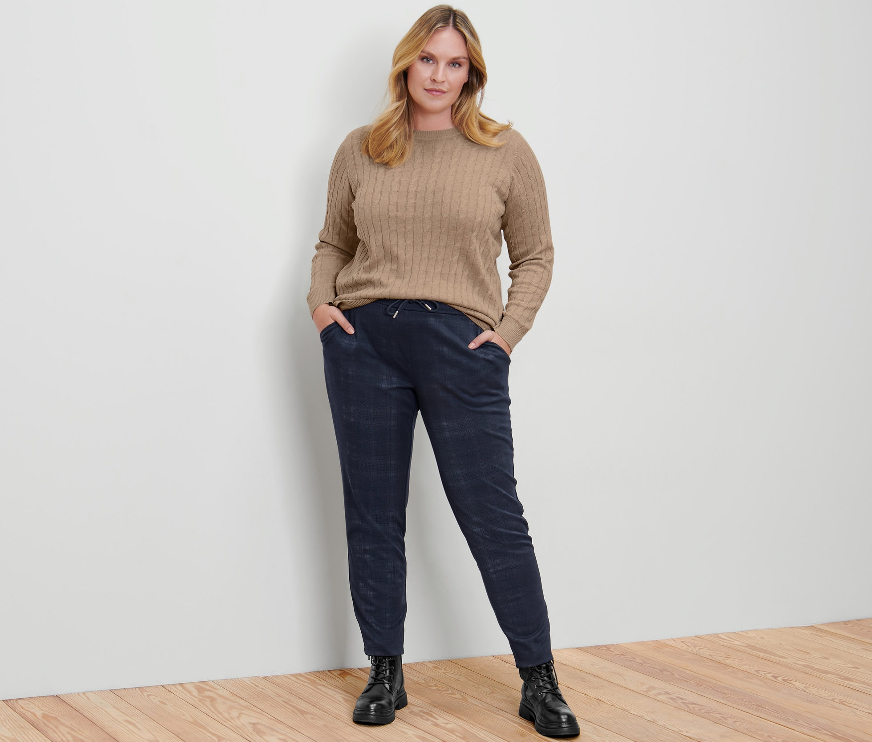 Frau posiert in Zopfstrickpullover, creme meliert, karierten Joggpants und Leder-Boots.