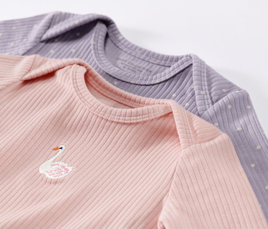 Nahaufnahme von zwei Baby-Langarmshirts mit Rippstruktur in Rosa und Lila. Das rosa Shirt hat eine Schwanenstickerei.