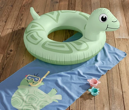 Ein grüner Schwimmring »Schildkröte« liegt auf einem blauen Handtuch mit Schildkrötenmuster und zwei Spielzeugen auf einem Holzboden.