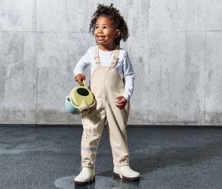 Mädchen in sandfarbener Kleinkinder-Regenhose hält eine Gießkanne, aus der Wasser läuft.