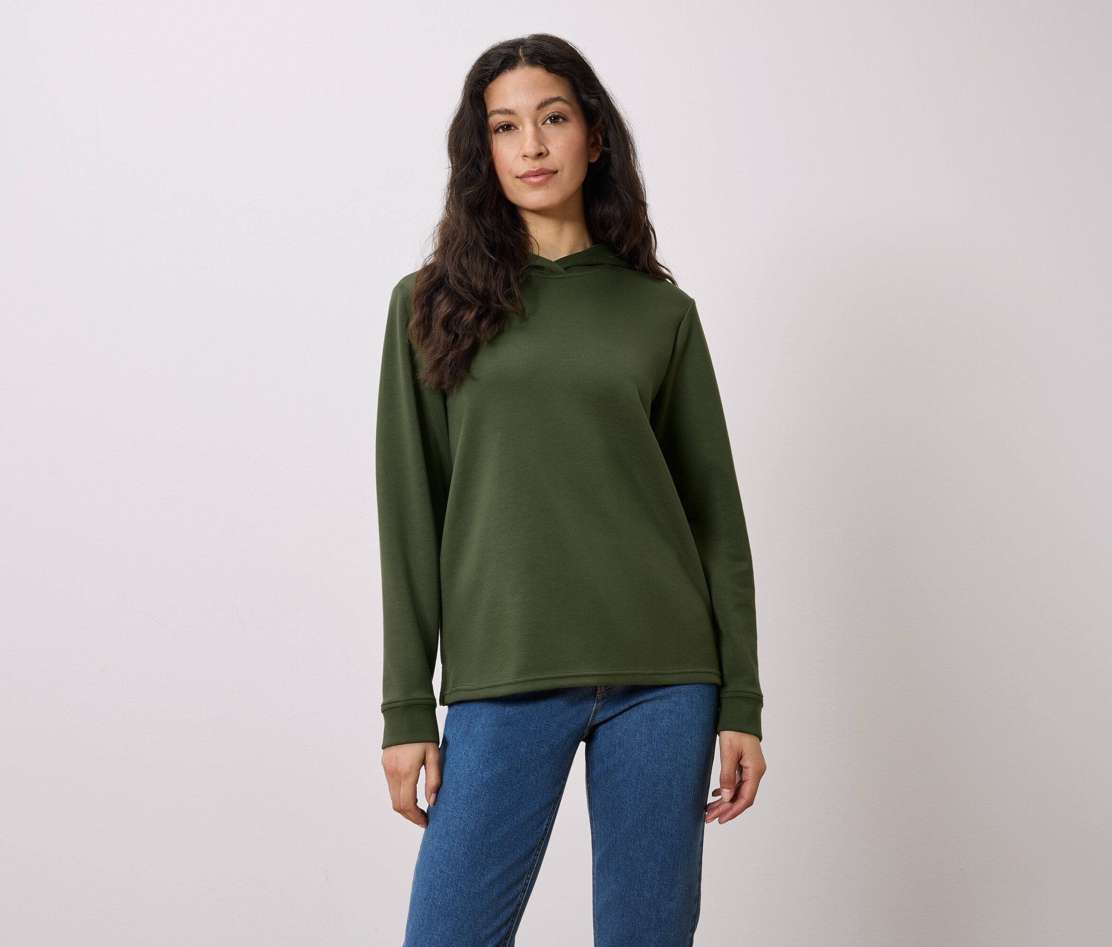Eine Frau mit langen braunen Haaren posiert in einem olivgrünen Hoodie und blauen Jeans vor einem weißen Hintergrund.