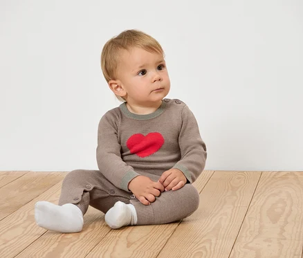 Baby sitzt auf einem Holzboden und trägt braune Kleidung mit einem Herz.