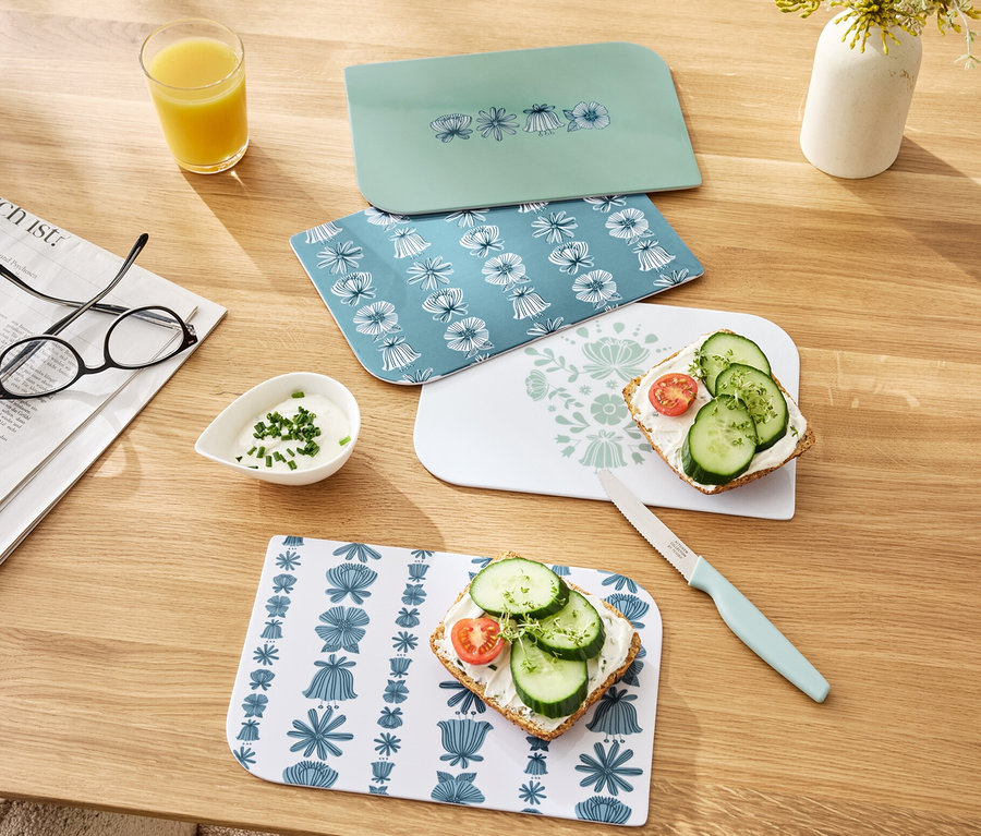 Vier Frühstücksbretter liegen auf einem Holztisch, zwei davon mit belegten Broten. Sichtbar sind auch ein Glas Orangensaft, eine Schale mit Dip und ein Messer.
