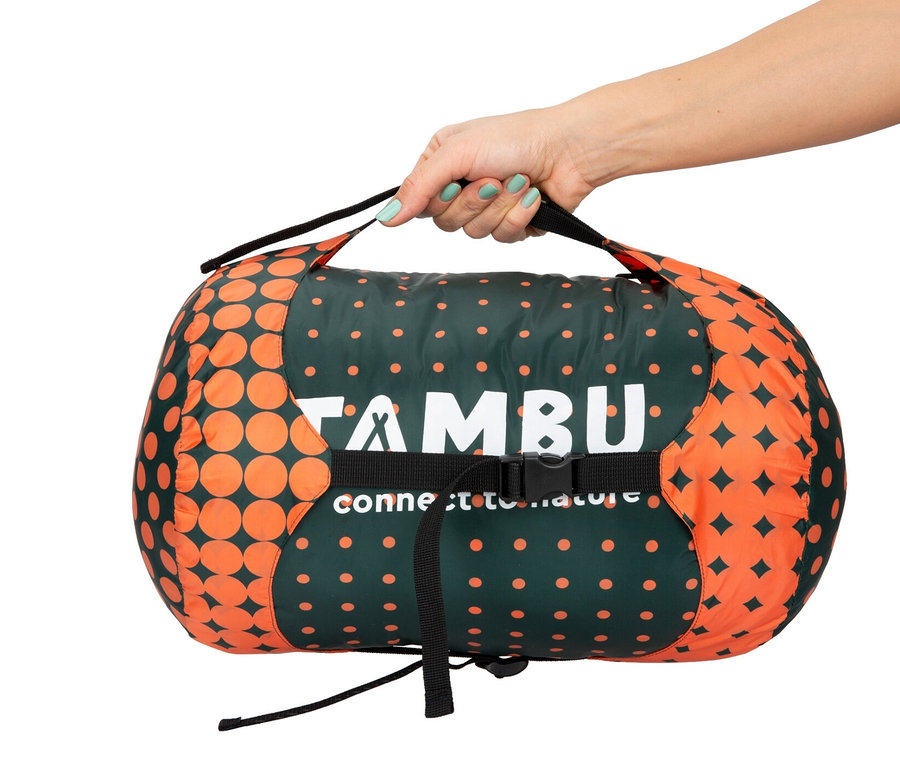 Eine Hand hält einen zusammengerollten Tambu Mumienschlafsack »KARA 1000«, der grün mit orangen Kreisen und Sternen ist.