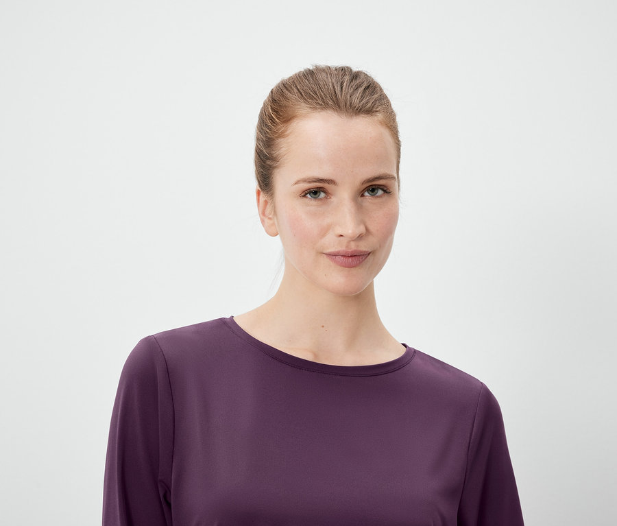 Porträt einer Frau mit zum Pferdeschwanz hochgebundenen Haaren, die ein lila Langarm-Oberteil vor einem weißen Hintergrund trägt.