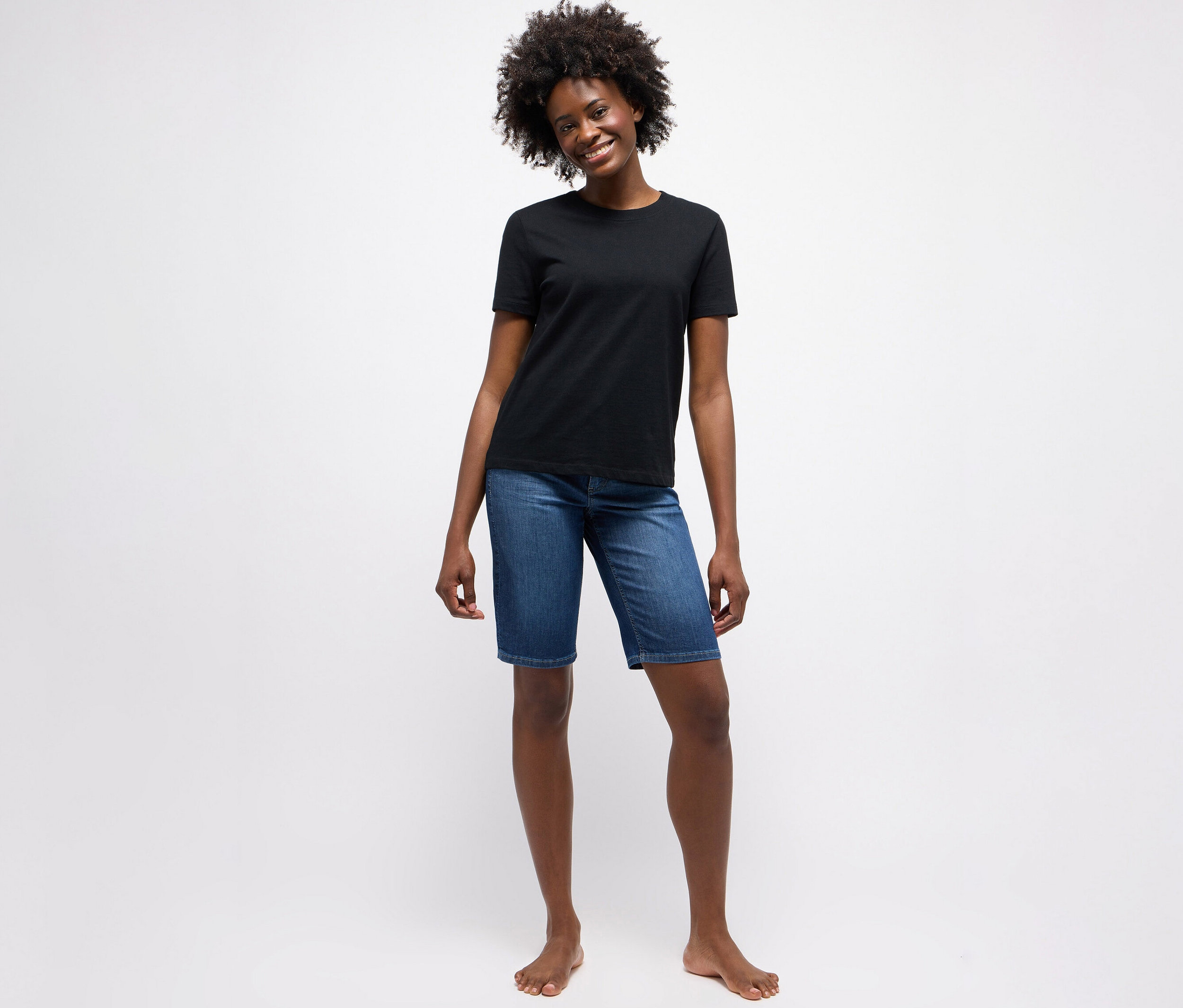 Eine Frau steht barfuß vor einem weißen Hintergrund und trägt ein schwarzes T-Shirt und eine "Angels Jeans »Bermuda Turn-Up«, Straight Fit, mid blue".