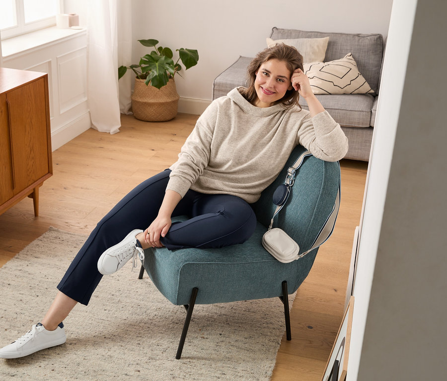 Eine Frau sitzt in einem Sessel und trägt einen Feinstrickpullover mit Kapuze, eine ausgestellte Leggings und Sneaker aus chromfrei gegerbtem Leder. Über den Sessel ist eine Crossbody-Tasche gehängt.